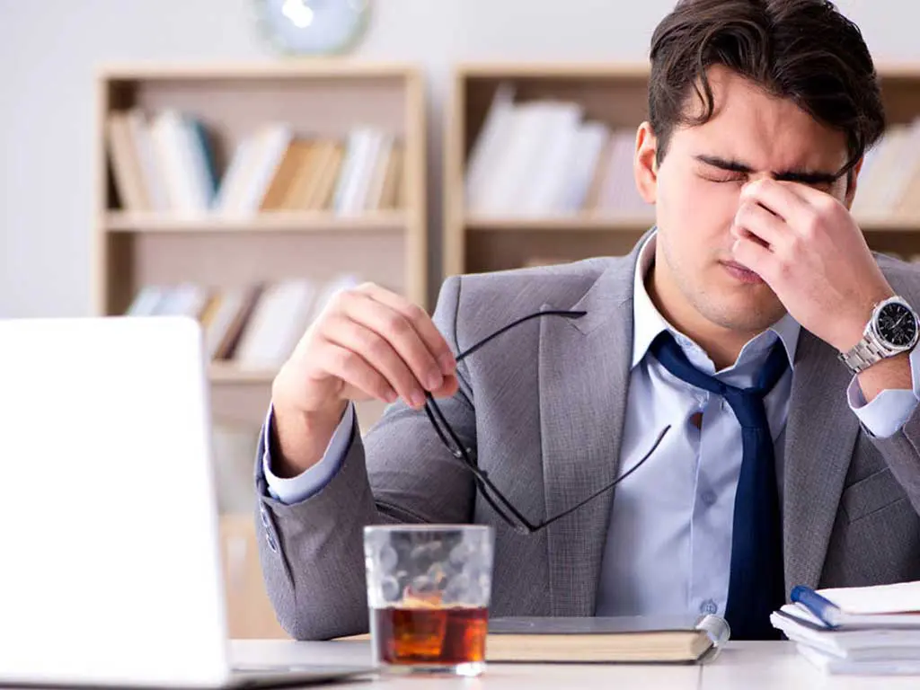A distressed-looking man in suit while there is a glass of alcohol at his desk