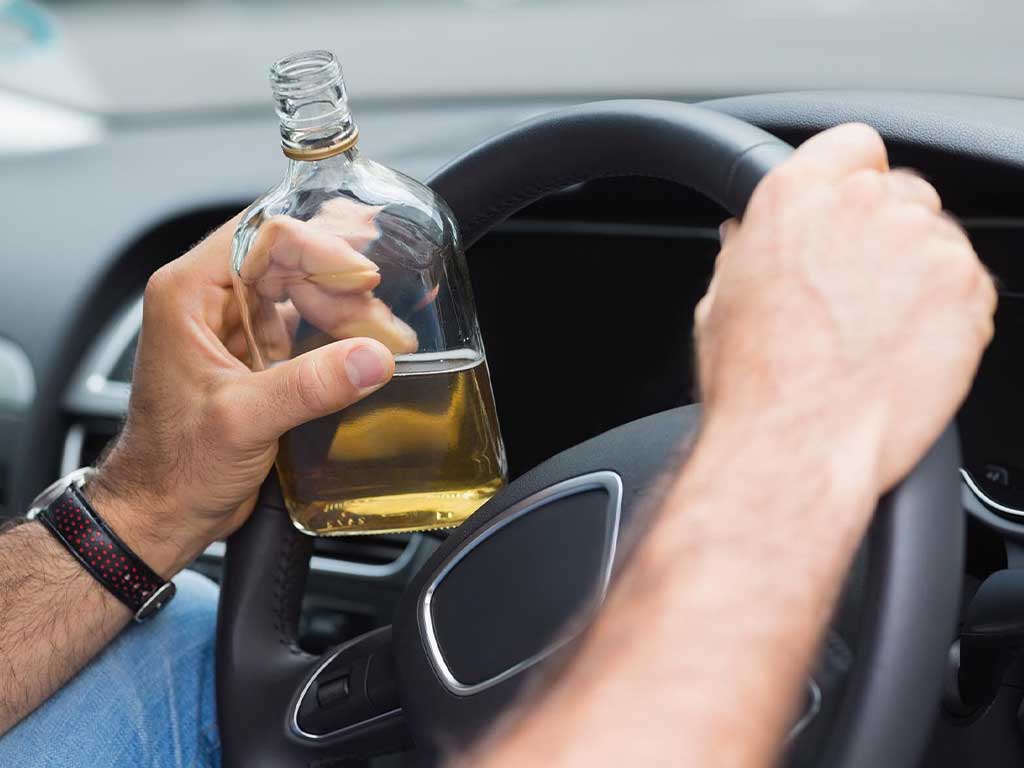 A person with an open bottle of alcohol holding the steering wheel